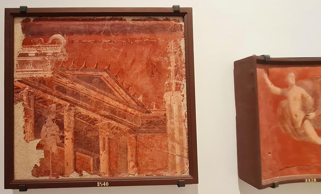 Herculaneum Augusteum. April 2023. Paintings on wall to left of Arch through to display of items from the Theatre.
On left – Building with tympanum on a red background – inv. 8540. Photo courtesy of Giuseppe Ciaramella.