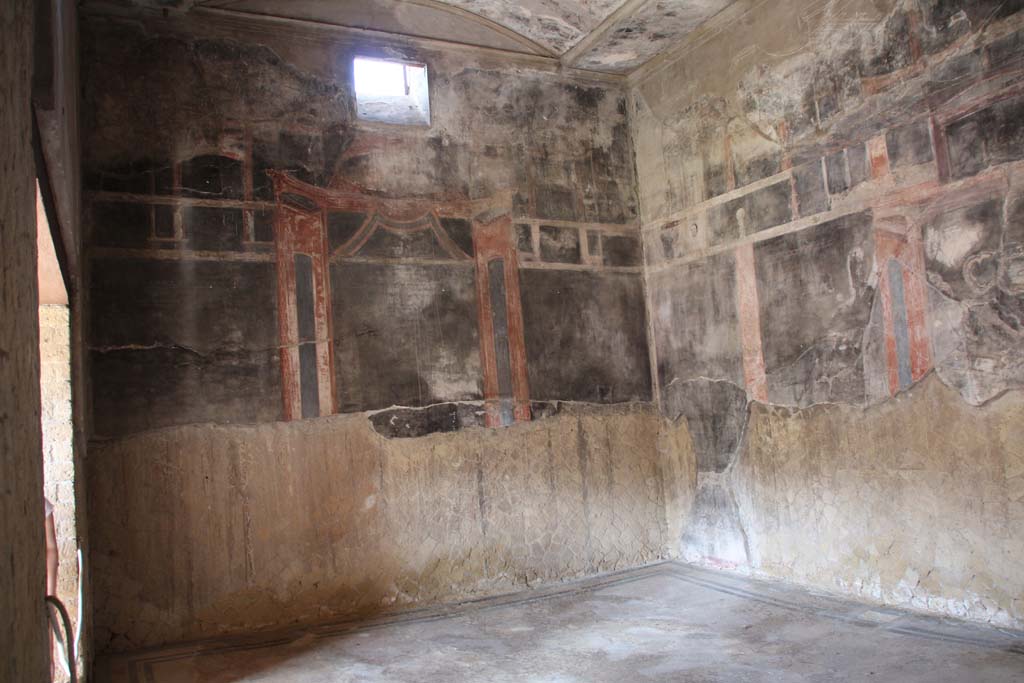 VI.13 Herculaneum, April 2014.
“Salone nero”, south wall with window, with entrance doorway (on left), looking towards south-west corner. Photo courtesy of Klaus Heese.
Maiuri wrote that on the western side (of the peristyle) is a large black hall, with the walls divided into three rectangular paintings like mirrors, flanked by pillars and chandeliers in lively colours.
The ceiling too, with a wide skylight (? vaulted area) in the centre, recomposed from various fragments, is on a black ground.
Against the wall is placed a wooden shrine in the usual form of a wooden temple, with wooden columns and little marble capitals, which preserved the little statues of a Lararium.
See Maiuri, Amedeo, (1977). Herculaneum. 7th English ed, of Guide books to the Museums Galleries and Monuments of Italy, No.53 (p.40).
According to Maiuri –
“Una mensa marmorea e un Larario in legno, appartenevano alle suppellettili di grandioso oecus.”
(A marble table and a wooden Lararium belonged to the furnishings of this grand oecus.)
See Maiuri A., 1958. Ercolano I Nuovi Scavi (1927-1958) Vol. 1. Roma: Istituto Poligrafico dello Stato, (p. 241).