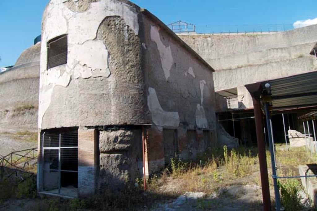 South-western baths, Herculaneum. July 2010. 
Looking towards baths complex 1, on left, separated by the adjacent terrace 2, with the lower level of large residential property, on right.
Photo courtesy of Michael Binns.
The three windows with their pyroclastic materials on the south side can be seen.
In the semi-circular apse, there would have been another three windows.
In the upper wall of the apse was a smaller rectangular window for light.
There were another three windows on the north side.
The terrace would have been bordered on the south and east by a portico, with flooring of cocciopesto with white tesserae dots.

