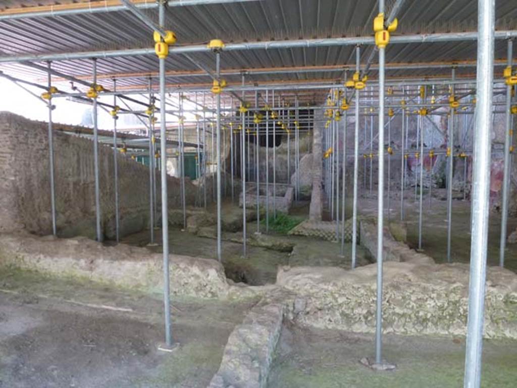 House of Dionysiac Reliefs, Herculaneum. June 2012. Looking north from room (f2), with room (q), lower left.
In the centre of the photo, on the left, is the biclinium room (n). Photo courtesy of Michael Binns.
Under the metal roof are the remains of the excavated area with fountain (p) and biclinium (n)
The biclinium, in poor state of preservation, preserves the preparation of a biclinium structure in masonry with rectangular basin in the corner.
On the west wall, besides the trace of a bourbon tunnel, one can observe the remains of the pictorial decoration in IV stile with grass tufts/plants.
See Guidobaldi, M.P, 2009: Ercolano, guida agli scavi. Naples, Electa Napoli, (p.132-3).